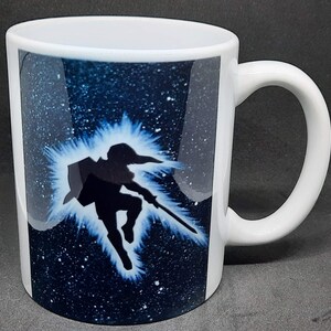 Può includere: Tazza da caffè in ceramica bianca con la silhouette di una figura con una spada su uno sfondo blu stellato. Il design è accentuato da un bagliore blu brillante. La tazza ha un manico curvo.
