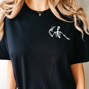 Op de afbeelding: Zwart T-shirt met een witte skeletprint die een martini-glas vasthoudt.