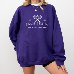 Puede incluir: Una sudadera morada oscura con el texto blanco "PALM BEACH POLO & RACQUET CLUB". El diseño incluye raquetas de tenis cruzadas y una pelota, con "EST 1928" encima del nombre del club. La sudadera es de estilo casual.