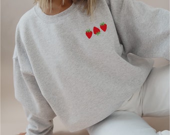 Peut inclure: Un sweat-shirt gris avec un petit motif brodé de trois fraises rouges sur le devant.