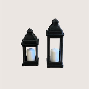 May include: Two black lanterns of different heights, each featuring a square frame with a pointed roof and a single white pillar candle placed inside the central compartment. The lanterns have a matte finish and a structured, geometric design.