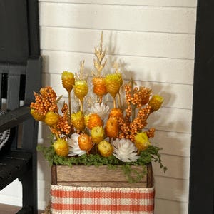 May include: A wooden crate filled with dried yellow and orange flowers, berries, and green moss. The crate is wrapped with an orange and white gingham ribbon and sits atop a square bale of straw, with a small pumpkin visible at the base.