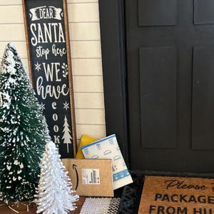 May include: A miniature Christmas scene with a black door, a sign that reads "Dear Santa stop here WE have COOKIES", and small decorative Christmas trees. Packages and a welcome mat are also present.