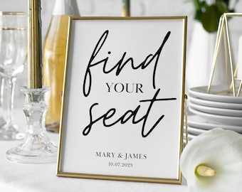 May include: Gold-framed sign with the text "find your seat" in black calligraphy. The sign is on a white tablecloth with a champagne bottle, glasses, and candles in the background. The text "MARY & JAMES 10.07.2023" is at the bottom of the sign.
