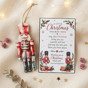 May include: A wooden nutcracker ornament with a red uniform and a jute string, paired with a rectangular card featuring the text A LITTLE CHRISTMAS POCKET HUG, may this Christmas bring you joy, warmth and love and may the new year bless you.