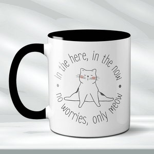Könnte beinhalten: Weiße Keramik-Kaffeetasse mit schwarzem Henkel und schwarzem Rand. Die Tasse zeigt eine Schwarzweißillustration einer Katze in einer Yogapose mit dem Text "in the here, in the now, no worries, only meow".
