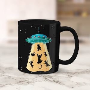 May include: Black coffee mug with a black and white illustration of a UFO abducting cats. The UFO is blue and green with a yellow beam of light. The cats are black silhouettes.