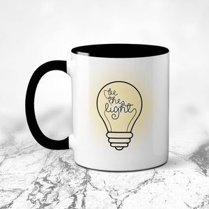 Könnte beinhalten: Weiße Kaffeetasse mit schwarzem Rand und Henkel. Die Tasse hat eine Glühbirnengrafik mit dem Text "be the light" in der Glühbirne.