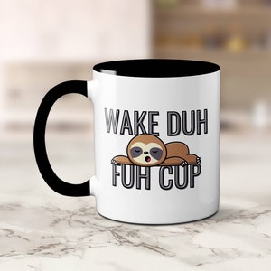 May include: White ceramic coffee mug with a black interior and handle. The mug features the text "WAKE DUH FUH CUP" with a cartoon sloth illustration. The sloth is brown and appears to be sleeping.