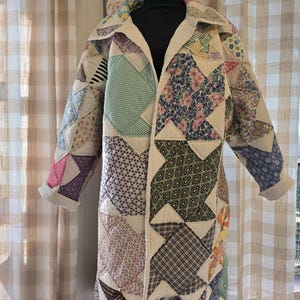 May include: A long-sleeved coat constructed from a patchwork quilt featuring a variety of colorful fabric squares in floral, plaid, striped, and geometric patterns. The garment has a wide collar, an open front, and visible white stitching.