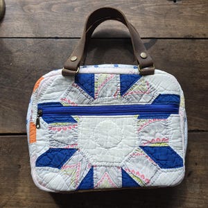 May include: A quilted handbag with a patchwork design in white, blue, and pink. The bag features a blue zipper and brown leather handles. The bag is approximately 10 inches wide and 7 inches tall.