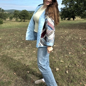 May include: A light blue quilted jacket featuring a patchwork sleeve with various plaid and patterned fabric strips, worn over a pale yellow top and light blue denim jeans, paired with beige slip-on shoes and white socks.