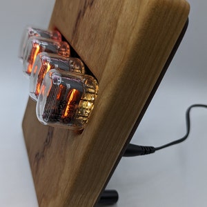 Könnte beinhalten: Eine Holzuhr mit einem schwarzen Kabel, das eingesteckt ist. Das Zifferblatt der Uhr besteht aus vier durchsichtigen Glasröhren mit orange leuchtenden Zahlen darin. Die Röhren sind mit den Nummern "MH226" und "CS9105" beschriftet.