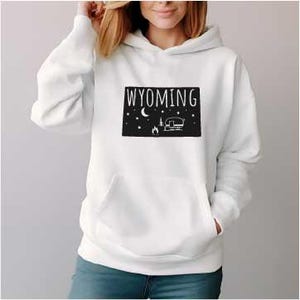 Può includere: Felpa bianca con un contorno nero dello stato del Wyoming, un camper, un falò, una luna e stelle. Il testo "WYOMING" è stampato in nero.