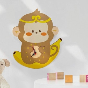 May include: A wooden wall clock shaped like a monkey sitting on a banana. The monkey has a yellow crown and a cute cartoon face. The clock face has black numbers and red hands.