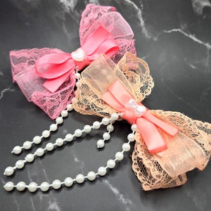 May include: Two decorative hair bows. One is pink with a pink satin bow and lace trim, the other is peach with a peach satin bow and lace trim. Both have a string of white pearls.