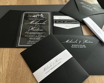 May include: A black wedding invitation suite featuring a clear acrylic card with white text, including the names Mikaela and Weston, a venue illustration, and date. The set includes black envelopes with white printed addresses and a white paper belly band.