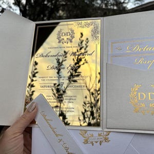 May include: Gold mirror wedding invitation with a white envelope and a white card with the text "Details" and "RSVP". The invitation features a gold wreath with the initials "DD" and the text "Together with their families, DeKendrick & DeShonda, request the honor of your presence at the celebration of their marriage, Saturday, 7 December, Two Thousand Twenty-Two, Time and Dreaming, One Hundred and One, Isla Vista Highway, Isla Vista, CA 93117, Please RSVP by November 1st, 2022, Directions to follow".