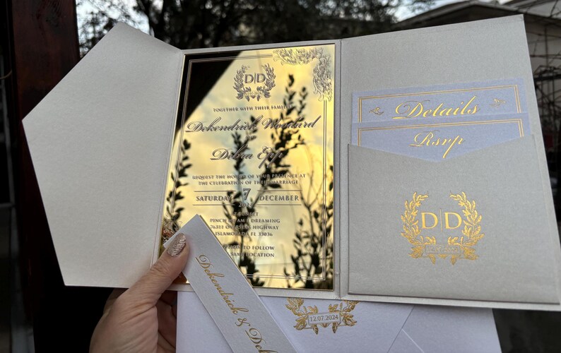 May include: Gold mirror wedding invitation with a white envelope and a white card with the text "Details" and "RSVP". The invitation features a gold wreath with the initials "DD" and the text "Together with their families, DeKendrick & DeShonda, request the honor of your presence at the celebration of their marriage, Saturday, 7 December, Two Thousand Twenty-Two, Time and Dreaming, One Hundred and One, Isla Vista Highway, Isla Vista, CA 93117, Please RSVP by November 1st, 2022, Directions to follow".