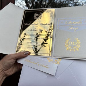 May include: Gold wedding invitation with a white background. The invitation features a gold mirror with the couple's names, "Dehondrick & Debra", and the date of the wedding. The invitation also includes a gold wreath with the initials "DD".