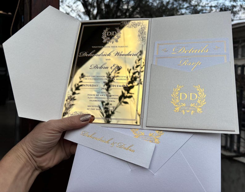 May include: Gold wedding invitation with a white background. The invitation features a gold mirror with the couple's names, "Dehondrick & Debra", and the date of the wedding. The invitation also includes a gold wreath with the initials "DD".