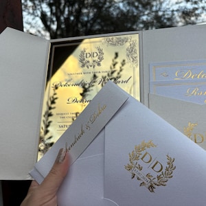 May include: A white wedding invitation with gold foil accents. The invitation features a gold mirror card with the couple's names and wedding date. The invitation also includes a white envelope with a gold foil monogram.