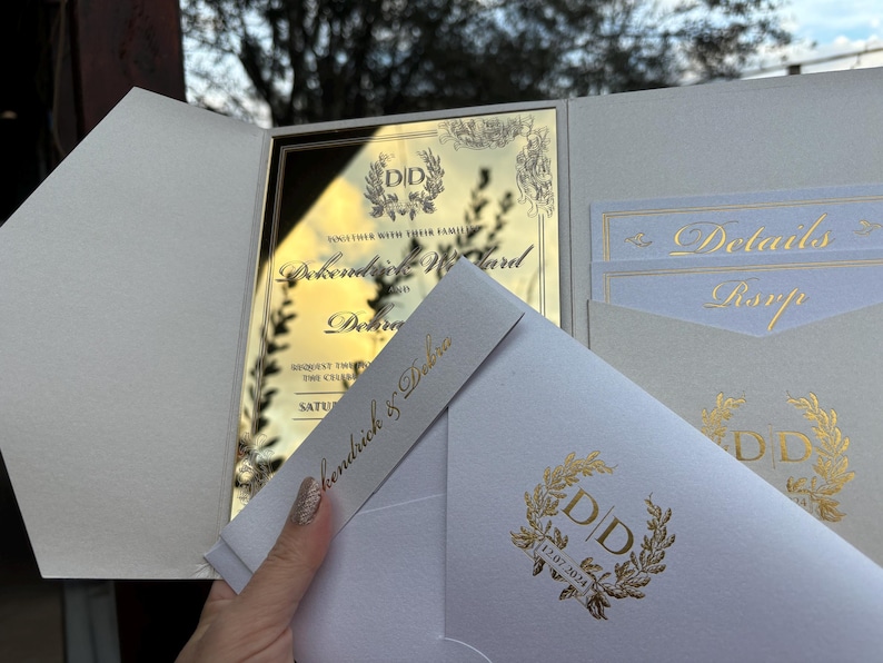 May include: A white wedding invitation with gold foil accents. The invitation features a gold mirror card with the couple's names and wedding date. The invitation also includes a white envelope with a gold foil monogram.
