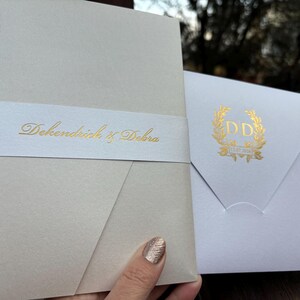 May include: Two white wedding invitations with gold lettering. The larger invitation has the text "DeKendrick & Debra" on a white band. The smaller invitation has a gold wreath with the initials "DD" in the center.