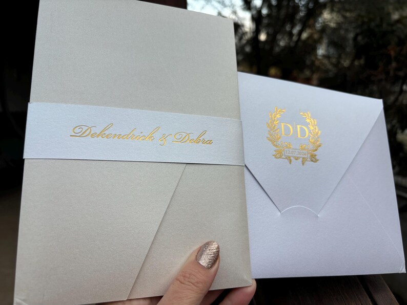 May include: Two white wedding invitations with gold lettering. The larger invitation has the text "DeKendrick & Debra" on a white band. The smaller invitation has a gold wreath with the initials "DD" in the center.