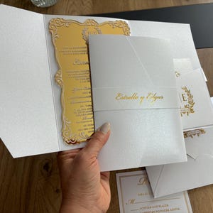 May include: Elegant white wedding invitation suite with gold accents. The suite includes a gold mirrored invitation card, a white envelope, and additional cards. The invitation features the names "Estrello y Edgar" in gold script.
