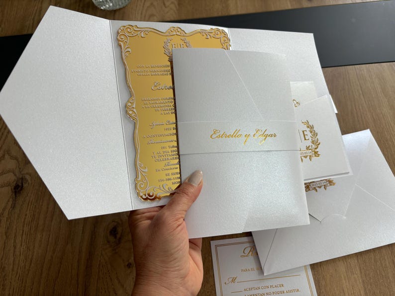 May include: Elegant white wedding invitation suite with gold accents. The suite includes a gold mirrored invitation card, a white envelope, and additional cards. The invitation features the names "Estrello y Edgar" in gold script.