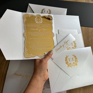 May include: White wedding invitations with gold accents. The invitation features a gold mirrored insert with the names "Estrella y Edgar" and decorative text. Matching envelopes and bands complete the set.