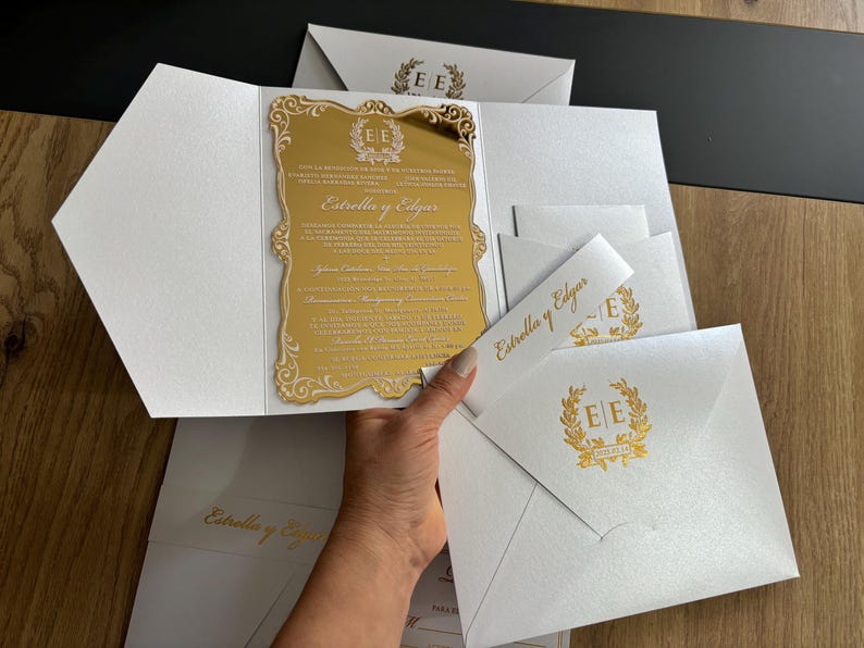 May include: White wedding invitations with gold accents. The invitation features a gold mirrored insert with the names "Estrella y Edgar" and decorative text. Matching envelopes and bands complete the set.