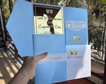 May include: A light blue wedding invitation with gold lettering. The invitation features the names "Lori K Donnelson" and "Clapton M Cota" and the text "Joyfully invite you to the celebration of their marriage".