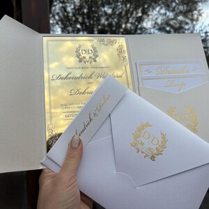 May include: A white wedding invitation with gold foil accents. The invitation features the couple's names, "Dokendrick & Dobra", and the date of the wedding. The invitation also includes a gold foil monogram with the initials "DD".