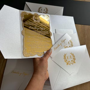 May include: Elegant wedding invitations featuring a gold mirrored insert with ornate detailing and the names "Estrella y Edgar." The invitations are presented in white, textured envelopes, with matching gold accents and text. Several envelopes are visible.