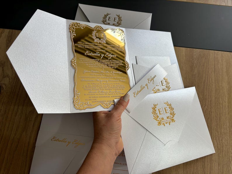 May include: Elegant wedding invitations featuring a gold mirrored insert with ornate detailing and the names "Estrella y Edgar." The invitations are presented in white, textured envelopes, with matching gold accents and text. Several envelopes are visible.