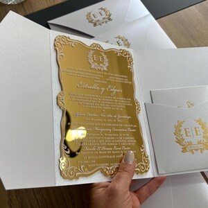 May include: A gold mirrored invitation with ornate edges and text, held open to reveal the details. The invitation is in a white envelope with a gold emblem. Additional white envelopes with gold emblems are visible.