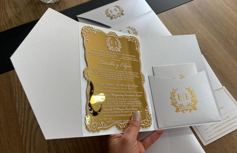 May include: A gold mirrored invitation with ornate edges and text, held open to reveal the details. The invitation is in a white envelope with a gold emblem. Additional white envelopes with gold emblems are visible.