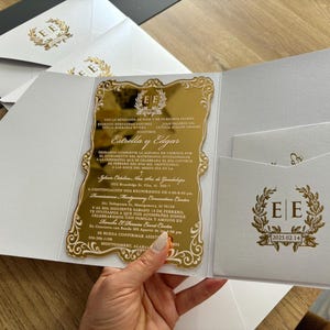May include: A gold mirrored invitation with ornate gold detailing and text, held open to reveal a white pocket with a matching gold emblem and text. Additional white envelopes with gold emblems are visible.