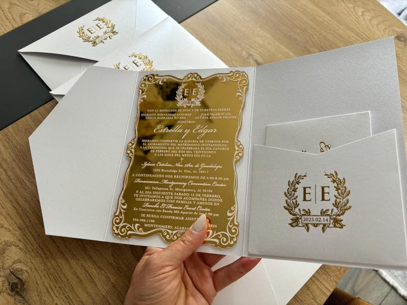 May include: A gold mirrored invitation with ornate gold detailing and text, held open to reveal a white pocket with a matching gold emblem and text. Additional white envelopes with gold emblems are visible.