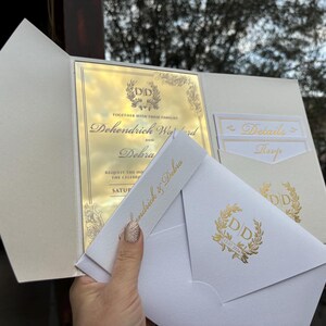 May include: A white wedding invitation with gold foil accents. The invitation features the couple's names, "Dehondrich Wynd and Debra", and the date of the wedding. The invitation also includes a gold foil monogram of the couple's initials, "DD", surrounded by a wreath.