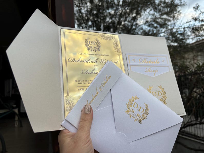 May include: A white wedding invitation with gold foil accents. The invitation features the couple's names, "Dehondrich Wynd and Debra", and the date of the wedding. The invitation also includes a gold foil monogram of the couple's initials, "DD", surrounded by a wreath.