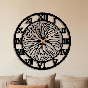 May include: A black, circular wall clock featuring Roman numerals and a tree-inspired design. The clock is mounted on a beige wall. The clock's hands are gold. The clock is above a white sofa with decorative pillows.