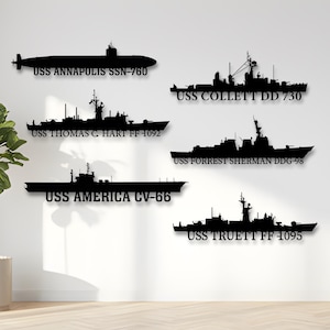 May include: Black metal wall art featuring silhouettes of U.S. Navy ships, including a submarine and various destroyers and cruisers. Each ship is labeled with its name and hull number, such as USS Annapolis SSN-760 and USS America CV-66. The art is mounted on a white wall.