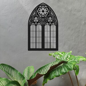 May include: A black metal gothic window wall art piece. The window features a pointed arch design with intricate tracery and a floral motif at the top. The window is mounted on a textured gray wall, with green plants in the foreground.