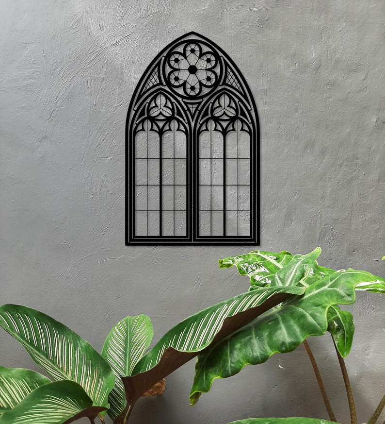 May include: A black metal gothic window wall art piece. The window features a pointed arch design with intricate tracery and a floral motif at the top. The window is mounted on a textured gray wall, with green plants in the foreground.