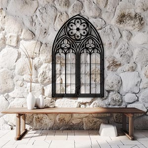 May include: Black metal gothic-style window frame mounted on a white stone wall. The window features an arched top with a floral design and two rectangular panes. A wooden table and white vases are in the foreground.