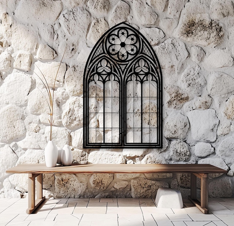 May include: Black metal gothic-style window frame mounted on a white stone wall. The window features an arched top with a floral design and two rectangular panes. A wooden table and white vases are in the foreground.