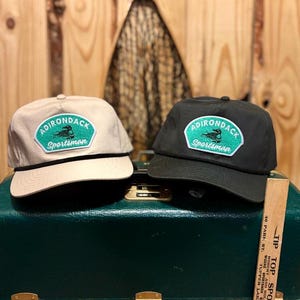 May include: Two baseball caps with a black and white patch that says "Adirondack Sportsman" with a green loon graphic. The cap on the left is beige and the cap on the right is black.